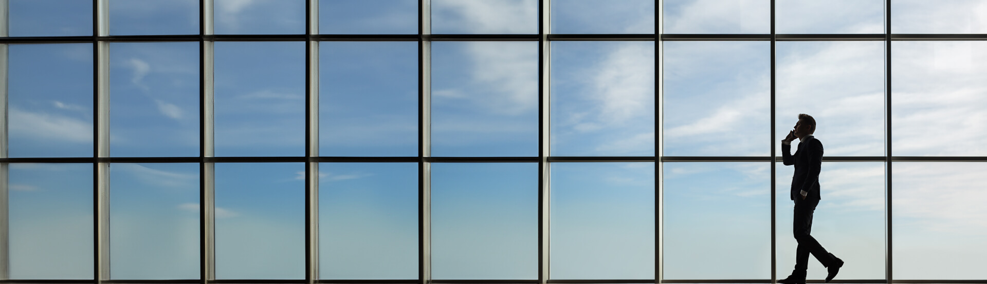 A man on a cell phone walking from right to left, the windows showing the sky in the background